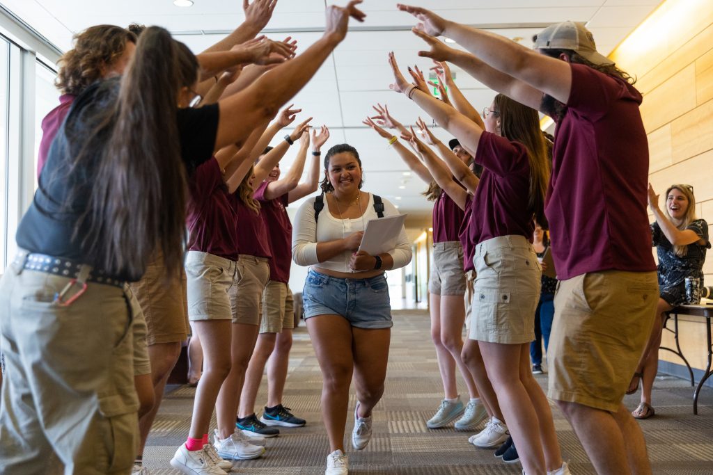 Jazzy Soria Gonzalez '24 walks through a cheering group of welcoming Valpo students during FOCUS, new-student orientation..Light blue and brown halftone graphic with radiating lines overlaying photo of Jazzy Soria Gonzalez '24 smiling at the camera in professional attire. 