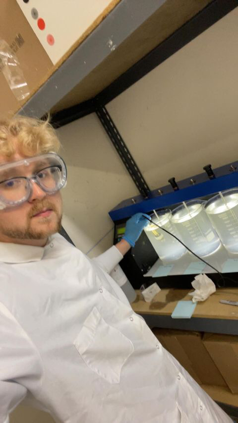 Daniel Owens '23 takes a selfie-style photo while interacting with lab equipment, wearing a white coat and goggles.