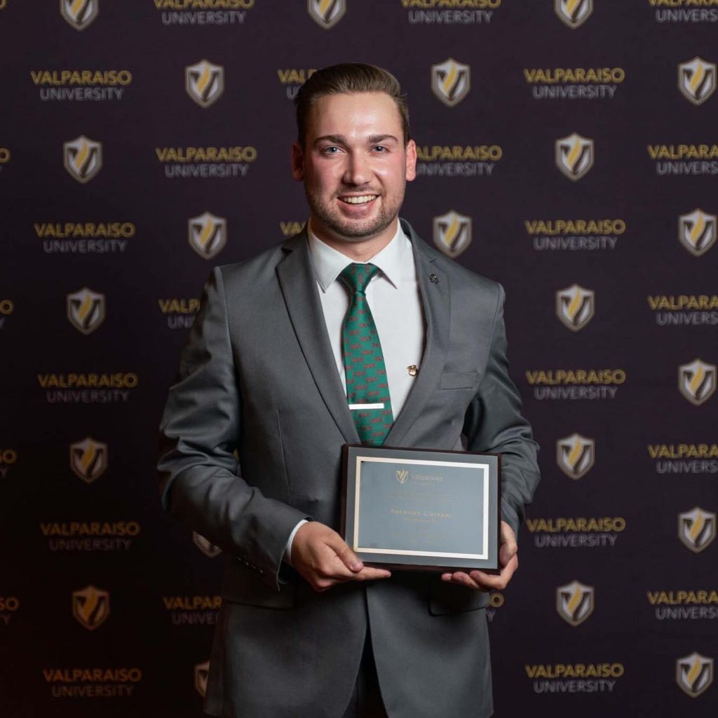 Valpo alumnus Anthony Luciano '22 smiles with a plaque in his hands in front of a University branded background and wearing professional attire.