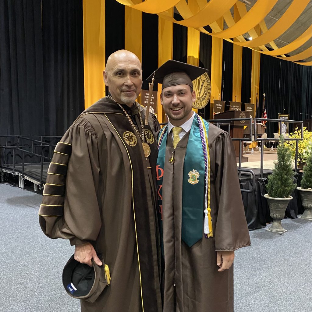 Valpo alumnus Anthony Luciano '22 smiles as he stands beside University President José Padilla, J.D., both wearing brown and gold Commencement regalia.
