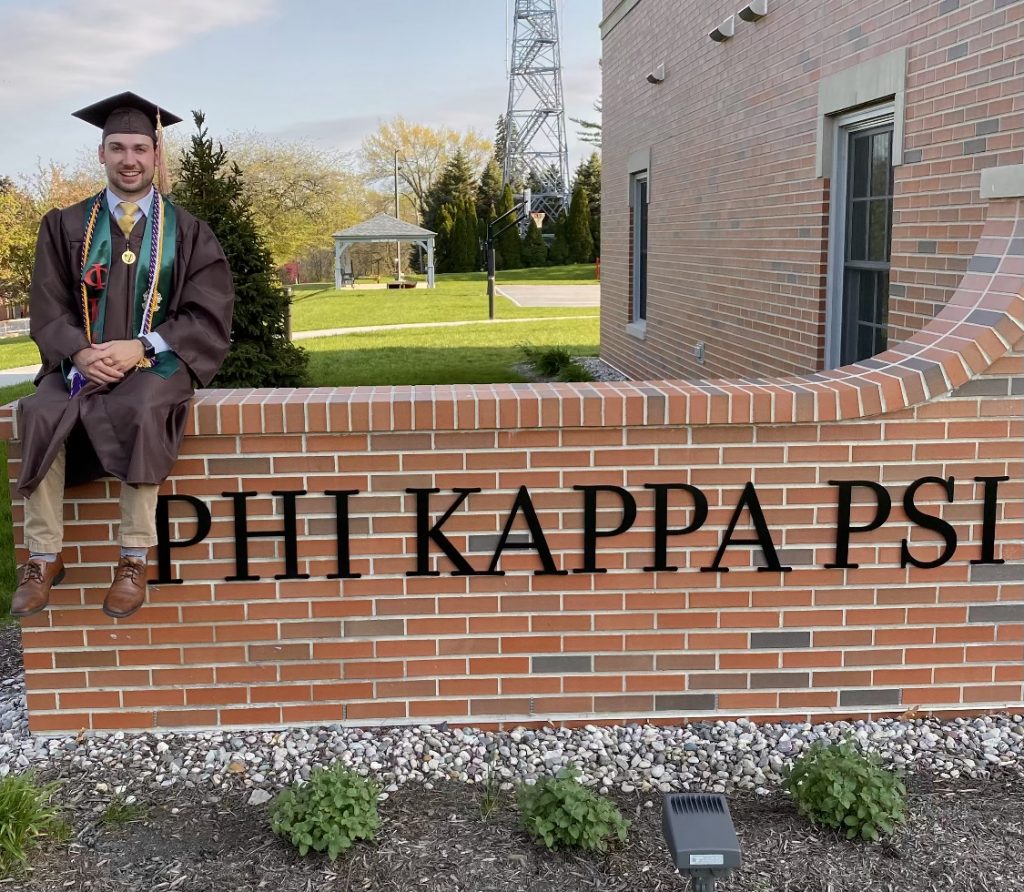 Valpo alumnus Anthony Luciano '22 smiles as he sits on top  of a brick partition that black letters reading, "Phi Kappa Psi" while wearing brown and gold Commencement regalia.
