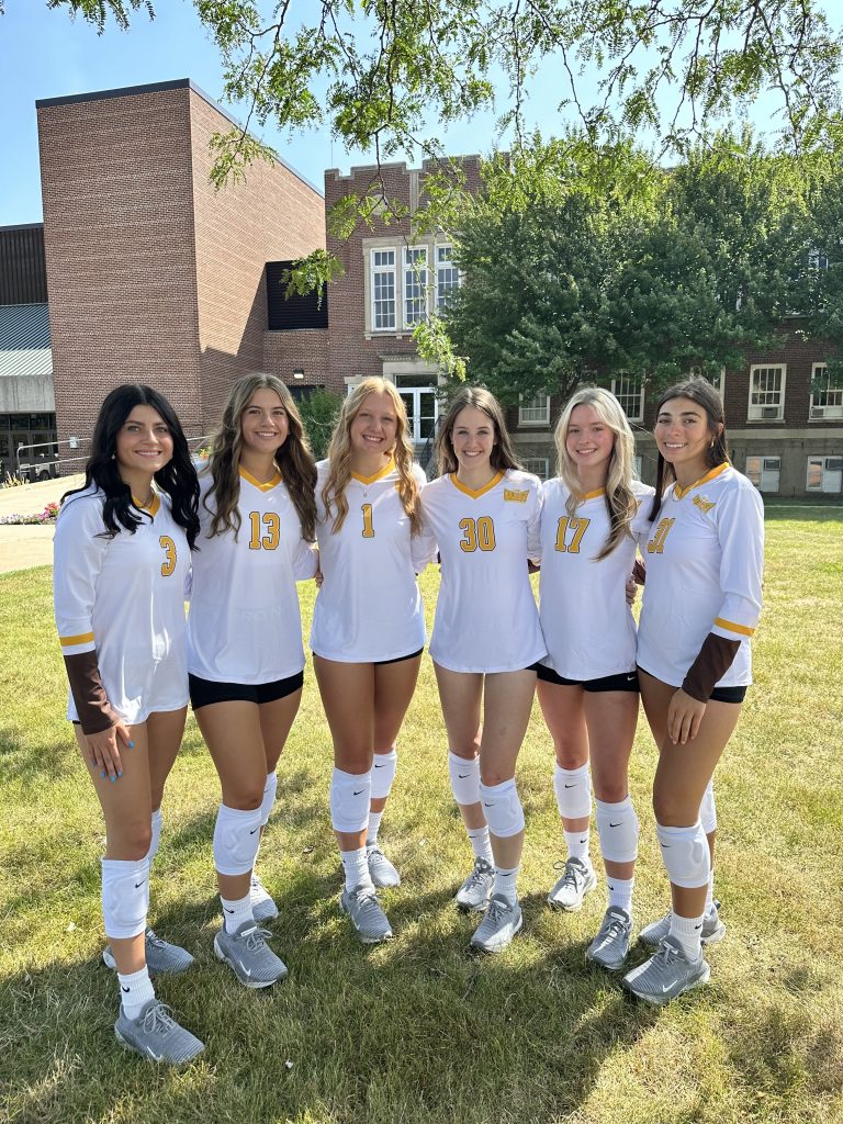 Valparaiso University student-athlete Emma Hickey '26 poses for a photo with her Valpo Volleyball teammates in front of the Athletics-Recreation Center (ARC) wearing matching white and gold uniforms.