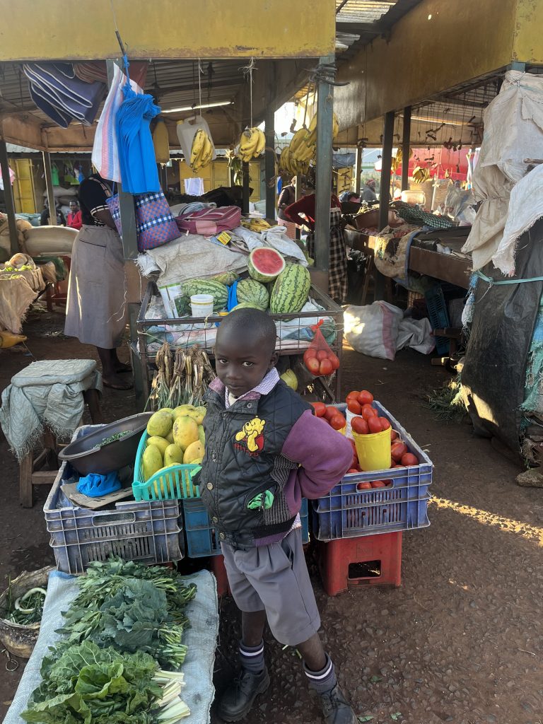 Young Kenyan child standing in front of a fruit cart and posing for a photo.