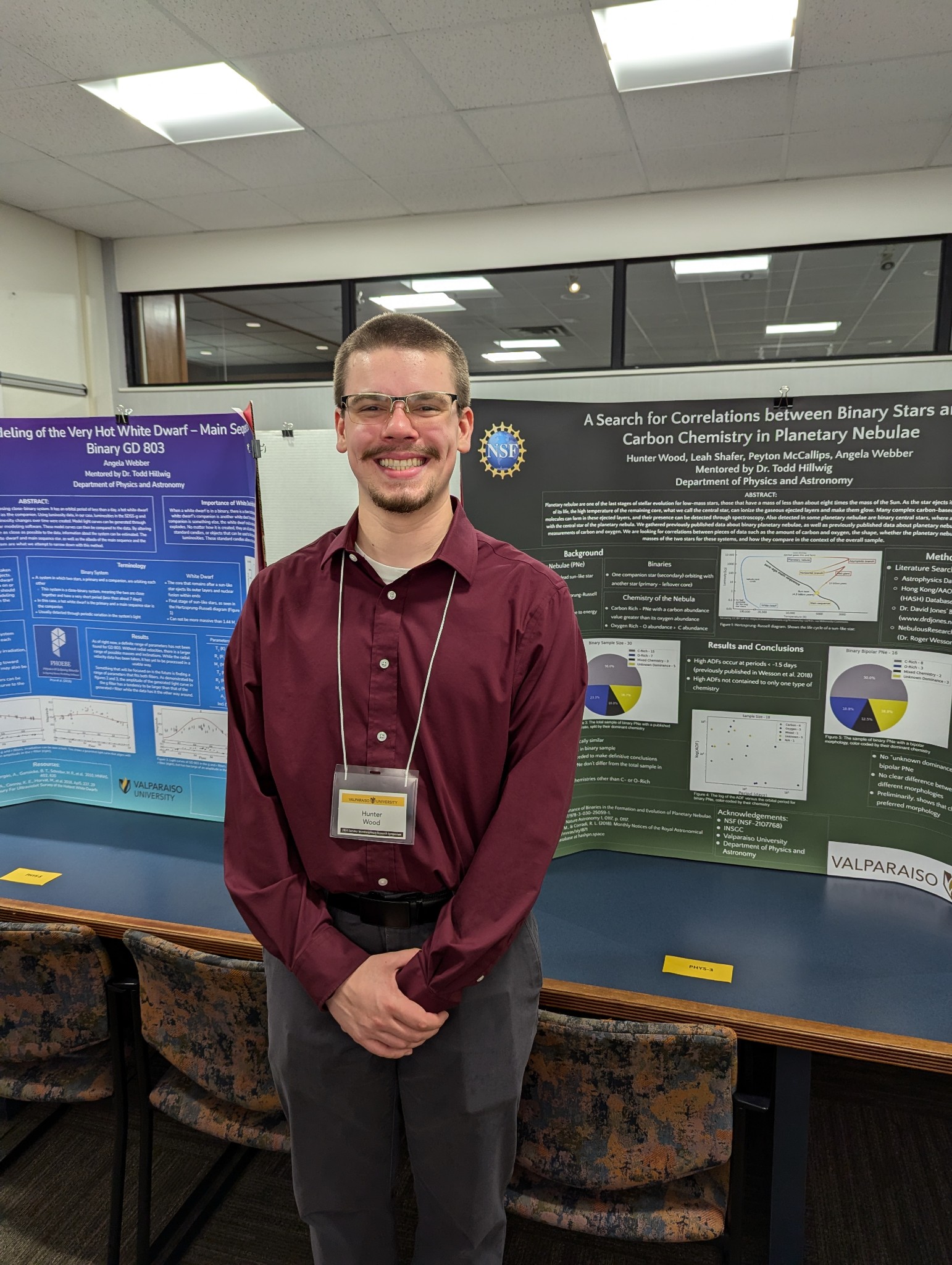 Valparaiso University astronomy student Hunter Wood '26 smiling as he stands in front of two poster boards filled with written and visual research.