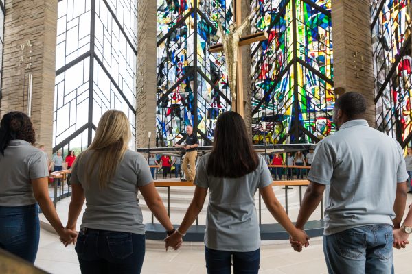 Four students stand hand-in-hand in front of the stained glass windows in the Chapel of the Resurrection