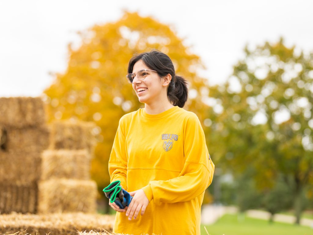 Student wearing a gold long sleeve shirt smiles away from the camera as she stands on campus.